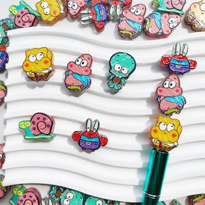 Könnte beinhalten: Eine Sammlung farbenfroher Cartoon-Charakter-Anhänger, darunter SpongeBob Schwammkopf, Patrick Star und Thaddäus Tentakel. Die Anhänger sind in verschiedenen Formen und Größen mit leuchtenden Farben und detaillierten Designs gehalten. Diese Anhänger sind wahrscheinlich für Schmuck- oder Bastelprojekte.