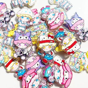 May include: A collection of colorful acrylic charms featuring cartoon characters. The charms showcase various designs, including characters with hats, bows, and outfits in shades of pink, blue, yellow, and purple. The charms are scattered across a white surface.