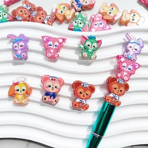 May include: A collection of colorful, cartoon character charms and a pen. The charms feature various animal designs in shades of pink, brown, green, yellow, and purple. The pen has a teal barrel and is topped with several brown bear charms.