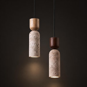 May include: Two pendant lights with a modern design. Each light features a cylindrical, porous, beige stone base topped with a wooden element. The lights hang from black cords against a dark background. The bottom of each light emits a warm glow.