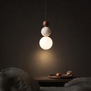 May include: A modern pendant light fixture with a unique design. It features a wooden sphere, a speckled stone sphere, and a large white globe light. The fixture hangs from a thin wire, creating a minimalist aesthetic. The light is on, illuminating the space below.