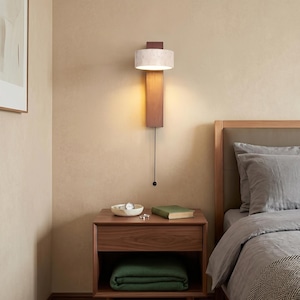 Könnte beinhalten: Eine moderne Wandleuchte mit rechteckiger Holzbasis und einem zylindrischen, marmorähnlichen Schirm. Die Leuchte hat eine Zugkette und ist an einer beigefarbenen Wand montiert. Darunter befindet sich ein Nachttisch aus Holz.
