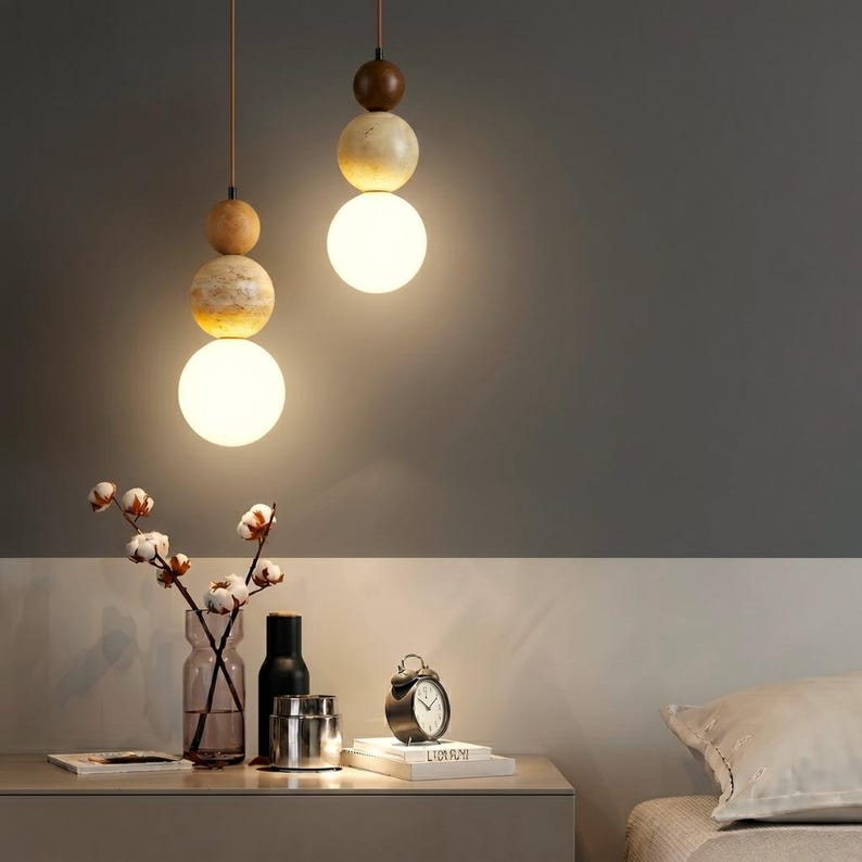 May include: Two pendant lights with a modern design. Each light features a series of wooden and marble-like spheres, topped with a round, glowing light. The lights hang from brown cords. The background is a neutral gray and white.