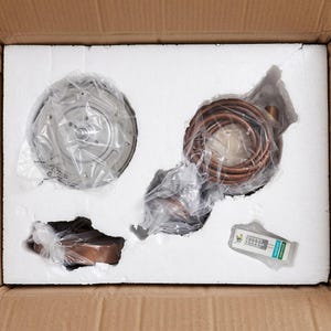 May include: An overhead view of a cardboard box containing HVAC components. The box is filled with white foam, and contains a circular metal component, copper tubing, and a small electronic device. The components are wrapped in plastic.