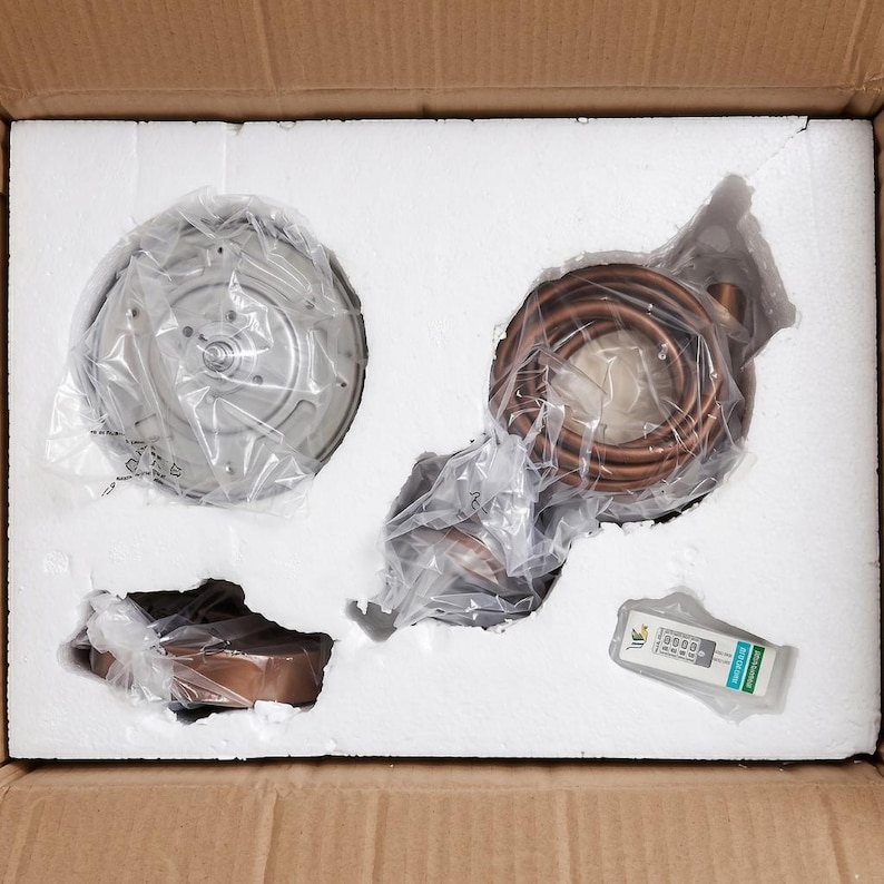 May include: An overhead view of a cardboard box containing HVAC components. The box is filled with white foam, and contains a circular metal component, copper tubing, and a small electronic device. The components are wrapped in plastic.
