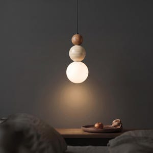 May include: A modern pendant light fixture with a stacked design. The fixture features a small, dark brown wooden sphere at the top, followed by a light-colored wooden sphere, and a large, glowing white glass sphere at the bottom. The light hangs from a black cord.