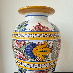 Op de afbeelding: Een keramieken vaas met een witte basis en een handgeschilderd bloemmotief in geel, blauw, groen en roze. De vaas heeft een brede gele rand, een taps toelopende hals en een afgerond lichaam met een craquelé glazuur over het oppervlak.