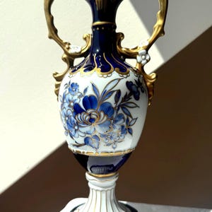 May include: A porcelain vase featuring a deep blue and white colour palette with hand-painted blue floral motifs and gold trim. The vessel has two ornate gold handles, a flared neck, and a pedestal base with a dark blue and white octagonal foot.