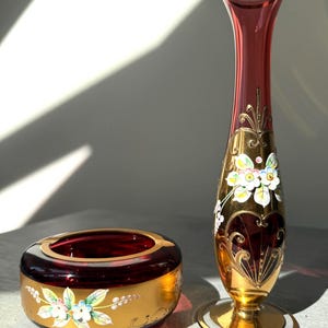 May include: A ruby red and gold glass vase and ashtray set. The vase is tall and slender with gold accents and floral designs. The ashtray is round with a gold rim and floral details. Both pieces have a vintage aesthetic.