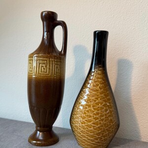 Op de afbeelding: Twee decoratieve keramische vazen. Eén is een hoge, bruine vaas met een handvat en geometrisch ontwerp. De andere is een druppelvormige vaas met een zwarte bovenkant en een getextureerd, amberkleurig lichaam. Beide vazen staan op een grijze ondergrond.