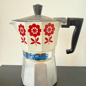 Könnte beinhalten: Ein vintage Espressokocher mit einem cremeweißen Oberteil und einem roten Blumenmuster. Er verfügt über einen schwarzen Griff und Deckelknopf, einen silberfarbenen Metallsockel sowie ein blaues Etikett mit der Aufschrift "MONEX VALTEX 35".