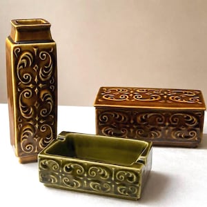 Può includere: Un set di tre oggetti in ceramica composto da un vaso rettangolare alto, una scatola rettangolare con coperchio e un posacenere rettangolare. Ogni pezzo presenta una smaltatura lucida marrone o verde oliva con un motivo in rilievo.
