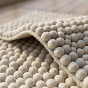 May include: Close-up of a beige rug with a textured surface. The rug is covered in small, round, raised balls, creating a 3D effect. The rug is folded over, revealing the underside.