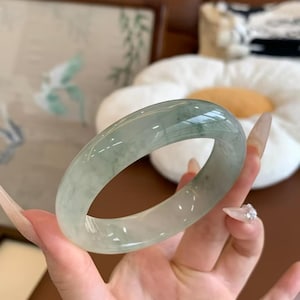 May include: A smooth, polished jade bangle bracelet featuring a translucent, pale green base with darker green mottled inclusions throughout the circular band. The stone has a glossy, reflective surface that catches the light, showcasing its natural mineral texture.