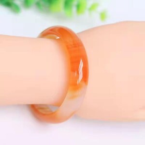 May include: A smooth, polished bangle bracelet crafted from translucent orange carnelian stone. The circular jewellery piece features natural colour variations ranging from light amber to deep reddish-orange tones with a glossy, reflective surface finish.