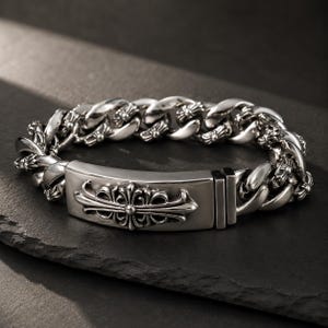 Op de afbeelding: Een armband van gepolijste zilveren schakels met een centrale rechthoekige plaat gegraveerd met een gedetailleerd, sierlijk kruismotief. De armband bestaat uit dikke, afgeronde schakels met kleine decoratieve kruisjes in de ketting.