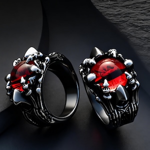 May include: Two black metal rings featuring a central, round, translucent red gemstone with a dark horizontal pupil-like slit. The band and setting are sculpted to resemble metallic claws or talons gripping the red stone securely in place.