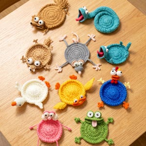 Könnte beinhalten: Eine Sammlung handgefertigter Häkeluntersetzer in verschiedenen skurrilen Tierdesigns. Die Untersetzer zeigen Kreaturen wie eine Maus, einen Frosch und einen Vogel in Braun-, Blau-, Gelb-, Rosa- und Grüntönen. Jeder Untersetzer hat einzigartige Details und Ausdrücke.