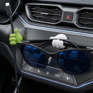 Peut inclure: Un support de câble en forme de pouce vert citron et un support de lunettes en forme de pouce blanc fixés sur un tableau de bord de voiture. Le support vert maintient un câble de charge noir, tandis que le blanc soutient des lunettes noires.