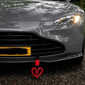 May include: A grey sports car featuring a black front grille, a yellow license plate, and a carbon fiber front splitter. A red heart-shaped tow strap with a broken heart design hangs from the lower front bumper area of the vehicle.