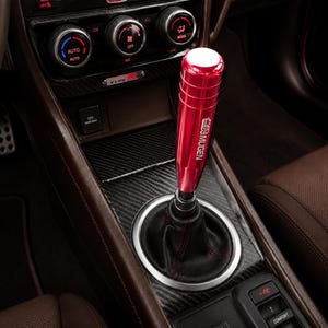 May include: A metallic red cylindrical gear shift knob with the white text MUGEN printed vertically on the side. The knob is attached to a black leather shift boot with red stitching, set within a carbon fiber textured center console of a vehicle.