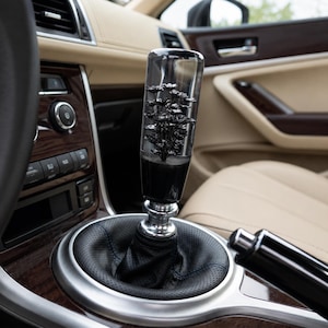 May include: A cylindrical gear shift knob made of clear resin containing a black floral arrangement inside. The knob is mounted on a silver metal base attached to a black perforated leather boot, positioned within a car center console.