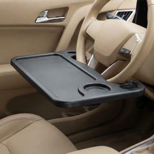 May include: A black plastic car steering wheel tray featuring a flat rectangular surface with a circular cup holder indentation and a smaller recessed area, mounted horizontally across the steering wheel of a vehicle with tan interior.