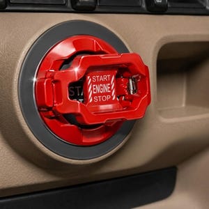 May include: A bright red, protective flip-up cover installed over a vehicle ignition button. The cover features a rectangular frame with a hinged mechanism and white text that reads START ENGINE STOP, with diagonal white stripes on the sides.