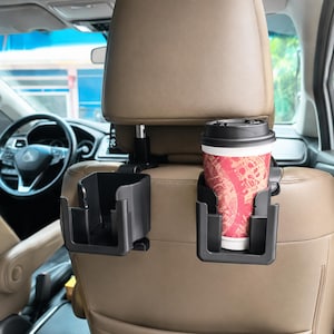 Puede incluir: Dos organizadores de asiento de coche de plástico negro fijados a los postes del reposacabezas. Un organizador está vacío, mientras que el otro sostiene un vaso de café de papel desechable con una funda estampada roja y blanca.