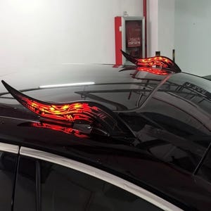 May include: A pair of curved, horn-shaped car roof attachments featuring a black base with intricate, glowing red tribal-style patterns. These decorative accessories are mounted on the glossy black roof of a vehicle, emitting a bright red light.
