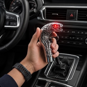 May include: A silver-colored gear shift knob shaped like a cobra snake head with a textured scale pattern and a glowing red LED eye. The metallic object is held in a vertical position above the center console of a vehicle interior.