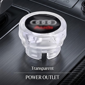 May include: A transparent plastic car power outlet cover featuring a circular black insert with the white Audi logo and red text. The cylindrical accessory has a notched base and a smooth, clear exterior finish designed for vehicle interiors.