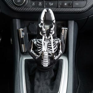 May include: A black and white miniature hoodie designed to cover a car gear shift knob. The fabric features a graphic print of a human skeleton rib cage and skull, complete with white drawstrings and metal aglets, fitted over a gear shifter.