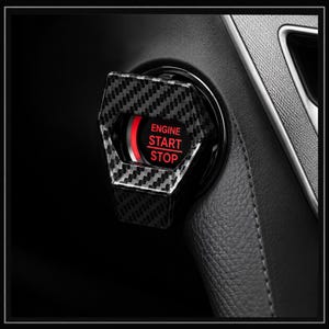 May include: A car engine start stop button cover featuring a black and gray carbon fiber pattern. The circular button is illuminated with a bright red light and displays the white text ENGINE START STOP centered within the circular frame.
