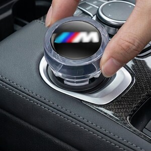 May include: A circular, clear plastic knob cover featuring the M logo with blue, purple, and red stripes on a black background. The accessory is designed to fit over a vehicle's central control dial, which is situated within a black leather console.