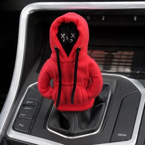 May include: A red fabric hoodie cover for a car gear shift knob. The hoodie features long black drawstrings and a small front pocket. Inside the hood, a black spherical head is visible, displaying two white X symbols for eyes and a small mouth.