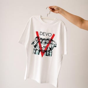 May include: White t-shirt featuring the band name "DEVO" in black letters above a red V-shaped design. Below the V is a graphic of five people. The shirt is held up by a hanger.