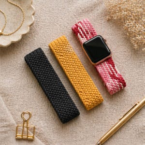 May include: Three knitted watch bands in black, mustard yellow, and a pink and red pattern. One band is attached to a black smartwatch with a gold case. The bands are lying on a beige surface with a gold pen and other accessories.