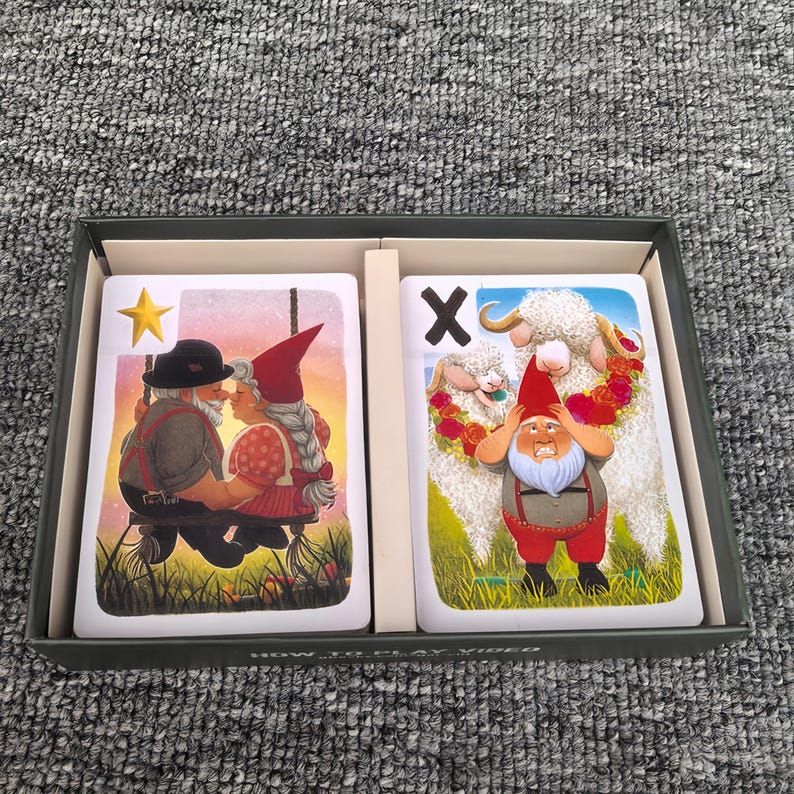 K&ouml;nnte beinhalten: Eine rechteckige Schachtel mit zwei Spielkarten. Die linke Karte zeigt einen gelben Stern und zwei Gnome auf einer Schaukel. Die rechte Karte zeigt ein schwarzes X und einen Gnom, der vor zwei wei&szlig;en Schafen mit Blumengirlanden steht.