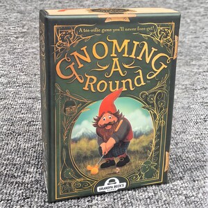 K&ouml;nnte beinhalten: Eine rechteckige Spieleschachtel f&uuml;r Gnoming A Round von Grandpa Beck's Games. Die dunkelgr&uuml;ne Schachtel zeigt goldene Zierr&auml;nder, eine Illustration eines Gnoms beim Golfspielen und den Text A tee-rific game you'll never fore-get! in Gold.
