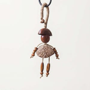 May include: A handcrafted wooden doll ornament with a mushroom-shaped head and skirt. The doll has wooden arms and legs, and is suspended by a beige rope. The ornament is designed with natural materials and textures.