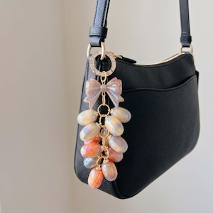 Op de afbeelding: Een zwarte leren handtas met een decoratieve sleutelhanger. De sleutelhanger heeft een doorschijnende roze strik en een cluster van iriserende, ovaalvormige kralen in tinten oranje, geel en blauw. De sleutelhanger is met een goudkleurige ring aan de tas bevestigd.