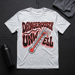 Op de afbeelding: Lichtgrijs t-shirt met de tekst "DANGEROUSLY UNWELL" in zwart en rood, met een thermometerafbeelding. De thermometer is rood en grijs, met een gebroken bovenkant en rode druppels.
