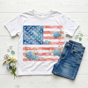 May include: A white short-sleeve t-shirt featuring a watercolor-style American flag graphic with blue hydrangea flower clusters integrated into the red and white stripes. The shirt is paired with blue denim shorts and a small bouquet of white and blue flowers.