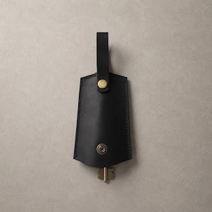 May include: A black leather key organizer featuring a trapezoidal shape with visible stitching along the edges. It includes a top strap secured by a brass-colored snap button and a lower brass-colored snap closure, with a metal key protruding from the bottom.