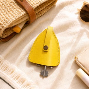 May include: A bright yellow leather key organizer with a triangular shape and a central snap button closure. The organizer features a metal key extending from the bottom, secured by a vertical strap that runs down the center of the yellow leather.
