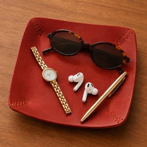May include: A square red leather valet tray with stitched corners containing a gold-tone wristwatch with a white face, a pair of white wireless earbuds, a gold-tone metal pen, and tortoise-shell patterned sunglasses with dark lenses.