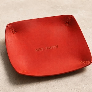 May include: A square red leather valet tray with rounded corners and visible cross-stitching at each corner. The centre of the tray is embossed with the text MRS. SMITH in a simple, capitalised serif font, creating a functional storage surface.