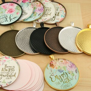 May include: Assortment of round metal pendant trays with a decorative swirl pattern. The trays are in various colors, including gold, silver, bronze, and black. There are also several round glass cabochons with floral designs and inspirational quotes, such as "Do small things with great love" and "There is always something to be grateful for".