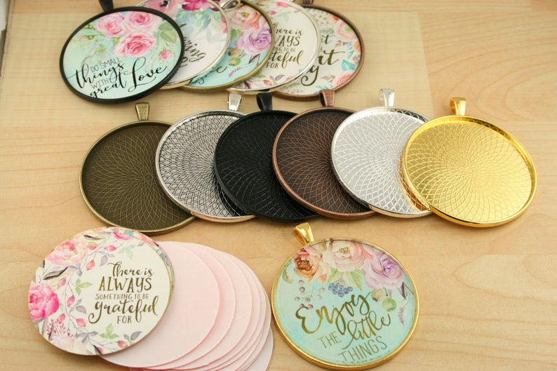 May include: Assortment of round metal pendant trays with a decorative swirl pattern. The trays are in various colors, including gold, silver, bronze, and black. There are also several round glass cabochons with floral designs and inspirational quotes, such as "Do small things with great love" and "There is always something to be grateful for".